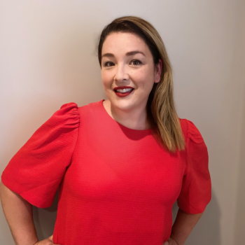 Smiling woman with shoulder length hair, wearing a red top standing against a plain light background.
